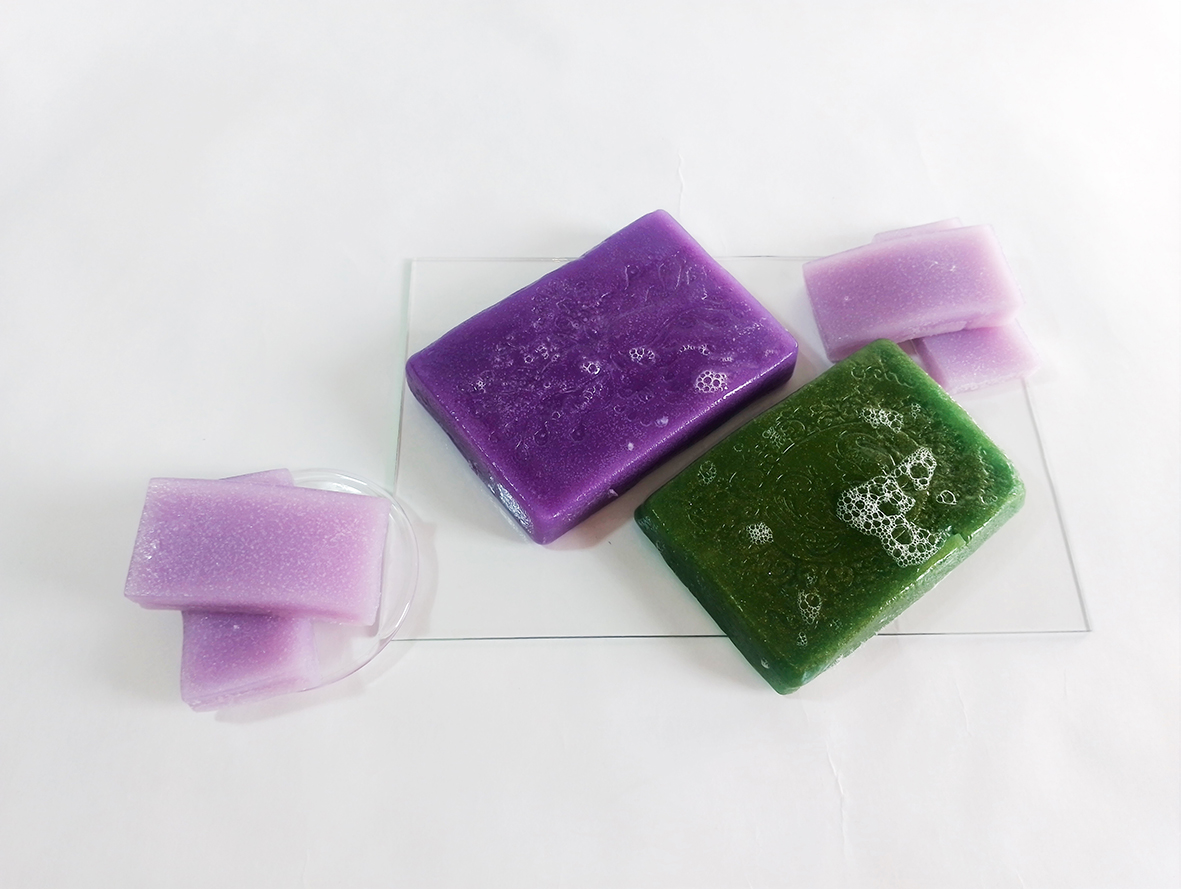 Sapone gelatina nei colori lavanda, viola e verde su una base acrilica trasparente posta su uno sfondo bianco.